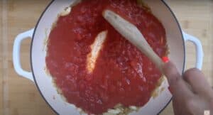 Stirring tomato sauce with a wooden spoon in a white pot on a wooden board background.